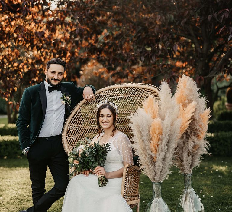 Bride sits in a peacock chair with pampas grass decor and the groom stands against the chair in a green velvet suit jacket 