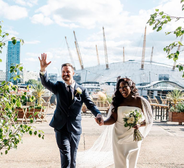 Black bride wears Suzanne Neville wedding dress and glitter polka dot tulle wedding cape as she walks with her groom in three-piece suit