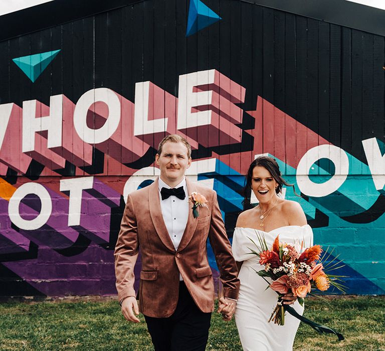 Bride in ASOS wedding dress and groom in pale pink velvet suit jacket with a colourful painted mural at The Wholehouse