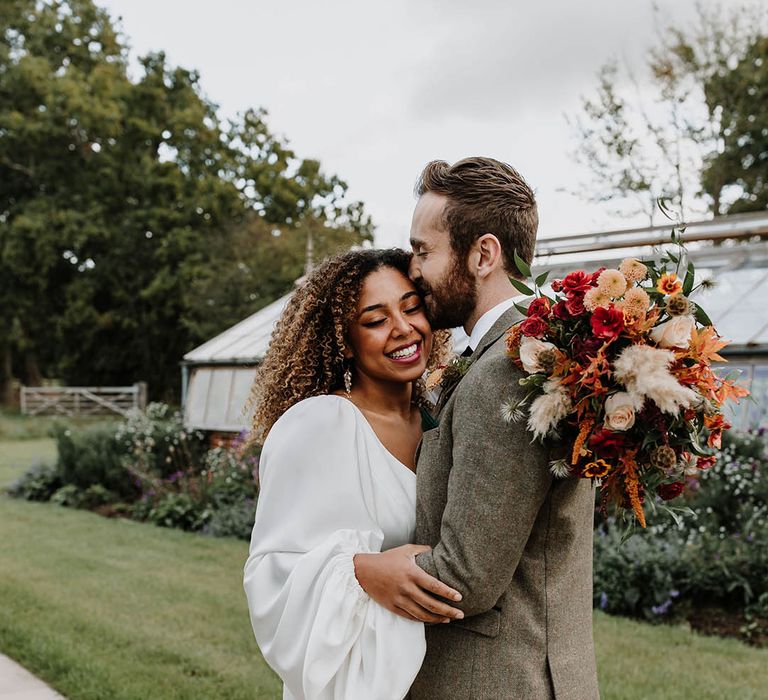 Groom in a grey suit kisses the bride on the forehead who wears a satin balloon sleeve wedding dress