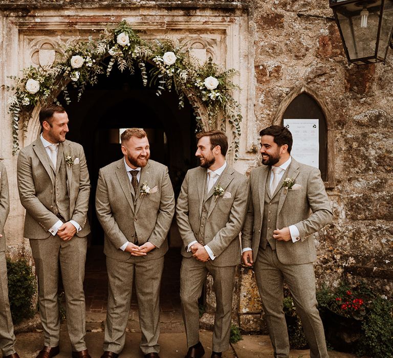 Groom and groomsmen laughing together in three piece grey suits with white flower buttonholes 