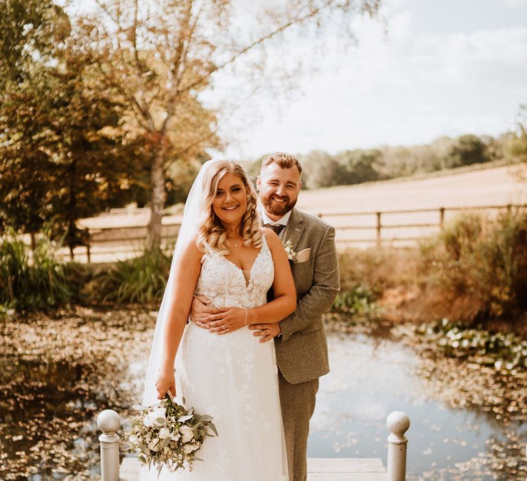 The groom in a three piece tweed suit embraces the bride from behind at the stunning location surrounding The Oak Barn