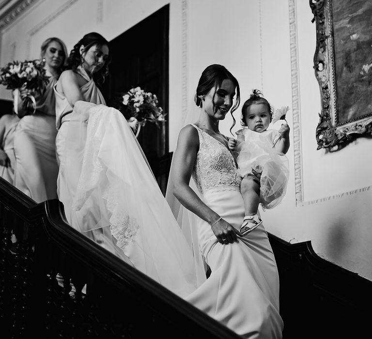 Bride walks down the stairs carrying her daughter followed by her bridesmaids