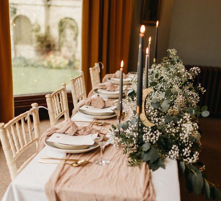 Neutral wedding tablescape with white table cloth, neutral pink table runner, sage green candles and foliage and baby's-breath table decorations