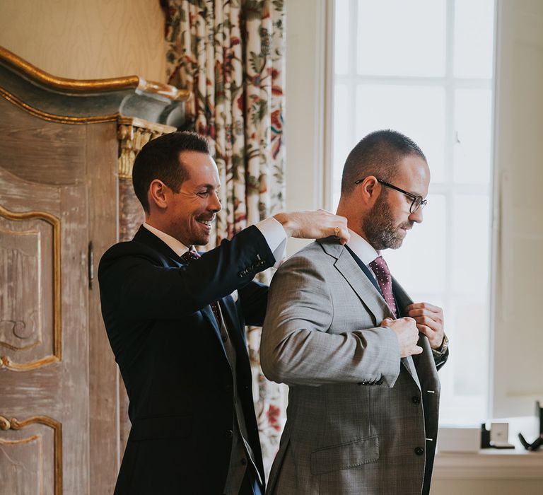 The groom in a blue suit helps the groom get into his light grey suit jacket for their wedding with deep red ties 