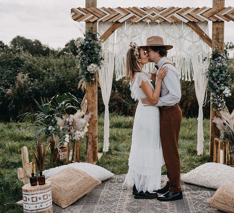 Bride in boho wedding dress with dried flower crown and groom in shirt and brown hat with macrame decor and boho styling 