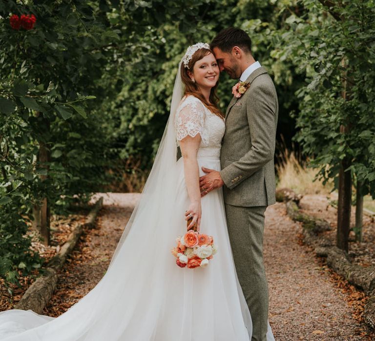 Bride wearing a lace glitter wedding dress with a princess skirt smiling at the camera being embraced by the groom in a tweed suit 