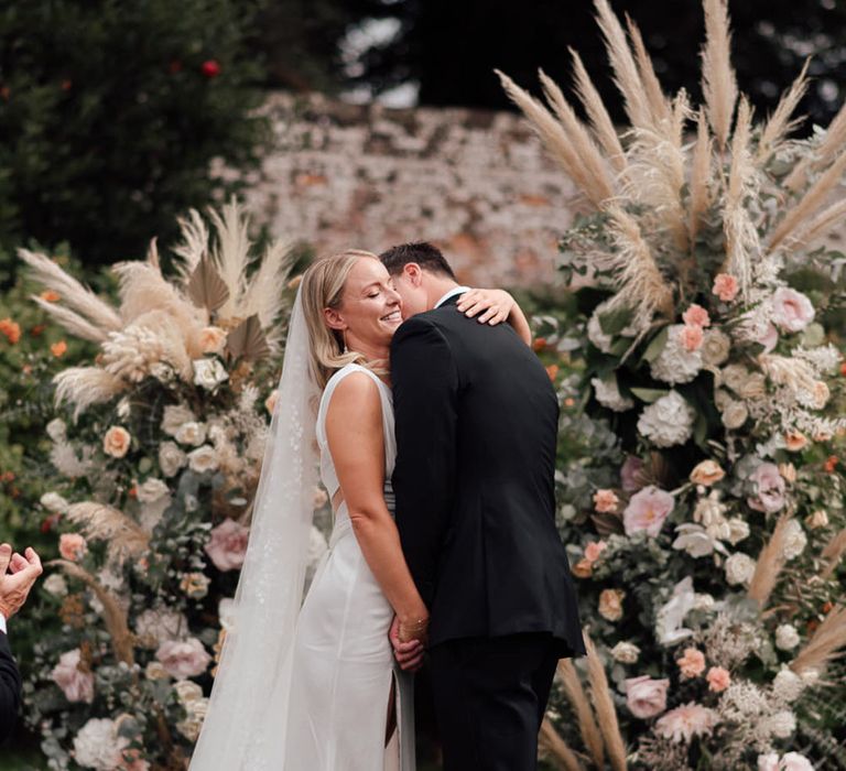 The groom kisses the bride on her neck as they stand at a the altar for their ceremony in front of large flower columns with pampas grass 