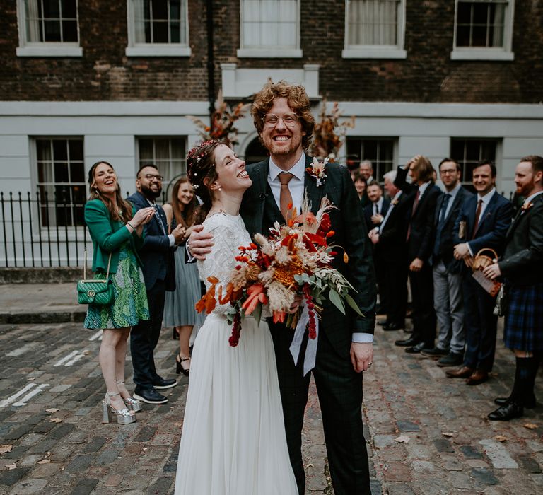 Groom in dark suit with orange tie and autumnal boutonniere embracing bride in long sleeve lace vintage wedding dress and closed toe satin heels holding large autumnal bouquet with orange, black, green and yellow roses, peonies, foliage and pampas grass surrounded by wedding guests on the street