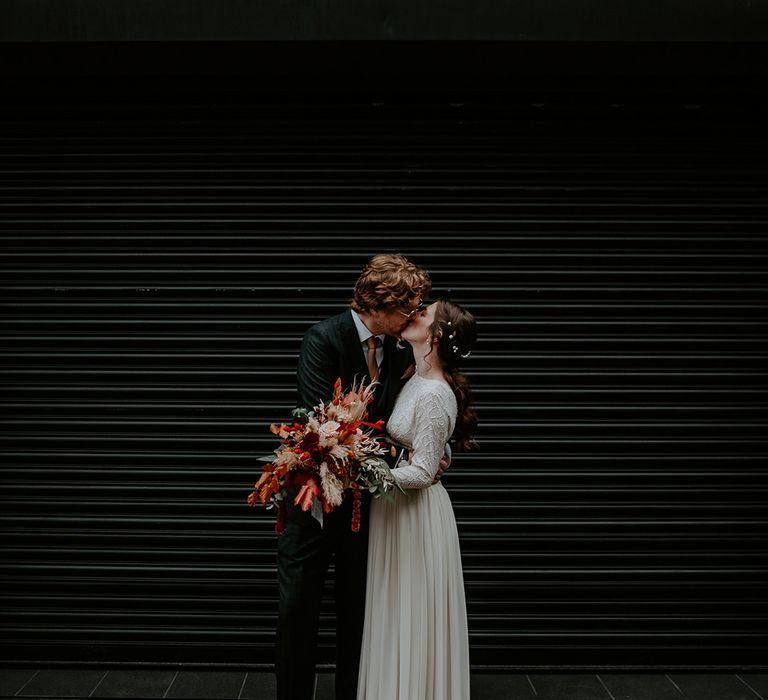 Groom in dark suit with orange tie and autumnal boutonniere kissing bride in long sleeve lace vintage wedding dress and closed toe satin heels holding large autumnal bouquet with orange, black, green and yellow roses, peonies, foliage and pampas grass