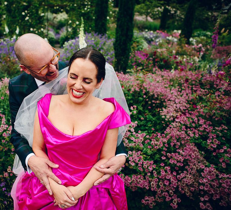 Groom in tan glasses and a dark forest green tartan suit embracing bride in off the shoulder front ruching pink wedding dress and two layer white and pink ombre veil