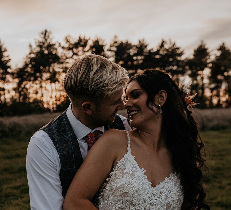 Bride in a detailed lace wedding dress with a flower pattern being embraced from behind with the groom in a grey waistcoat and paisley orange tie