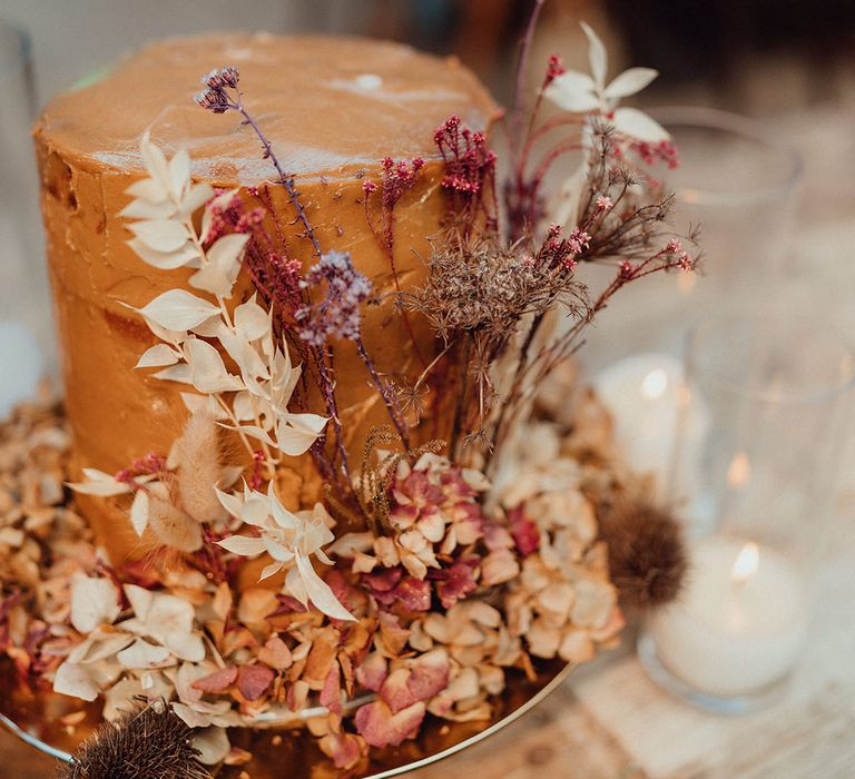 Tall orange frosted wedding cake with dried flower decoration for boho wedding next to pillar candles 