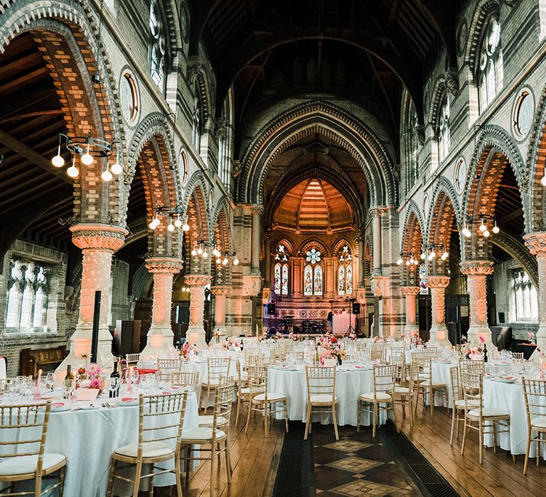 Round tables covered in white tablecloths complete with colourful floral arrangements and pink stationery surrounded by fairy lights wrapped around church pillars