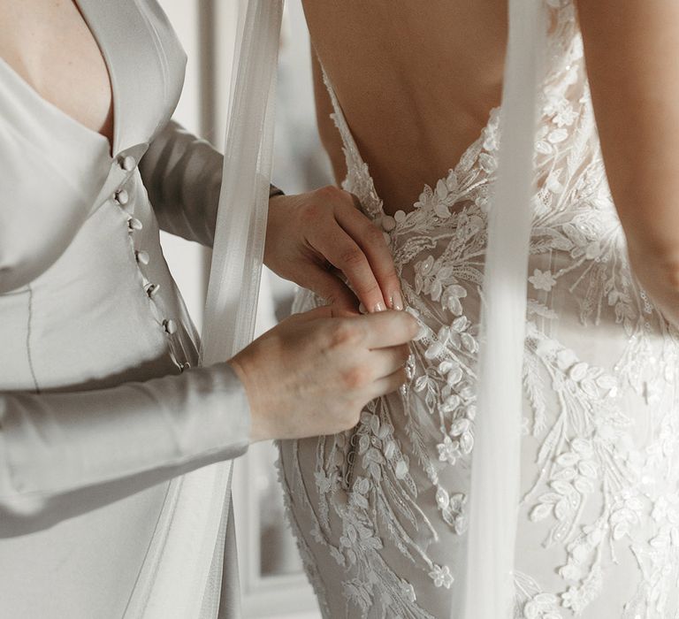 Open back flower sparkly patterned wedding dress worn by the bride being buttoned up by bridesmaid 