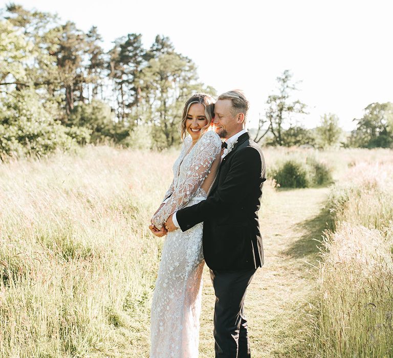 Groom in black tie embracing the bride from behind wearing a 3D lace wedding dress with an open back design