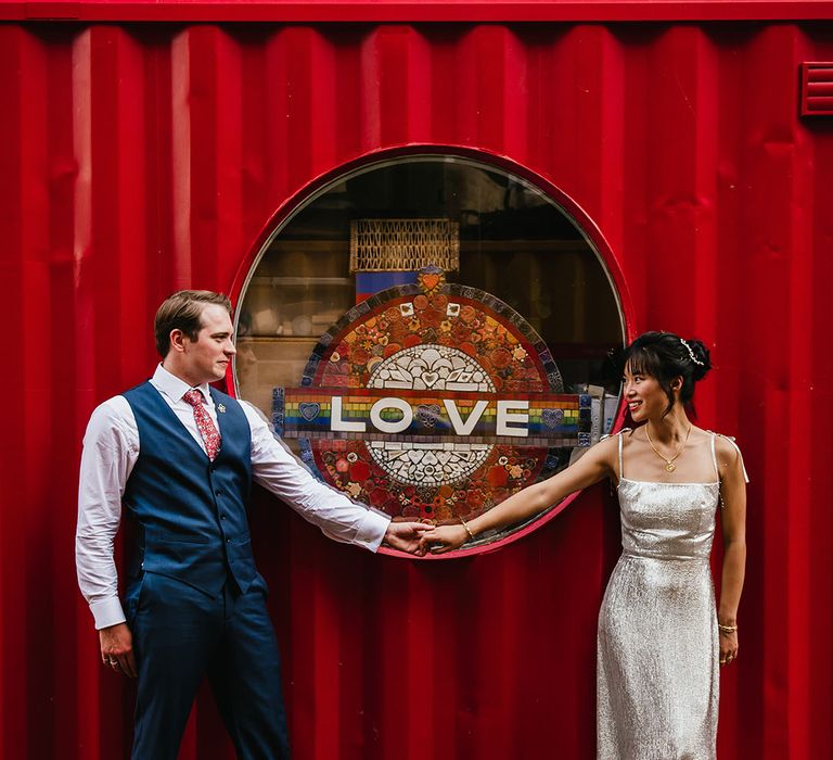 Bride & groom stand in front of red backdrop complete with round window and Love sign 