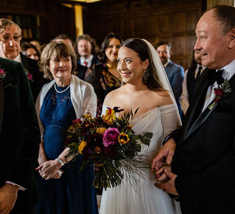 Father of the bride in traditional black tie walks the bride down the aisle to the groom at the end of the altar the bride holds a purple and yellow spring bouquet 