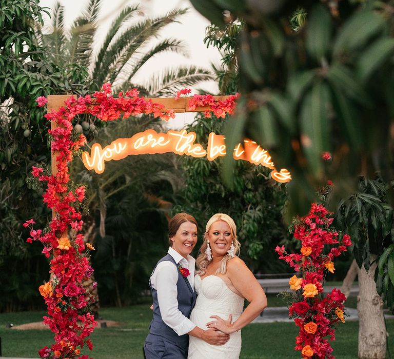 Brides stand beneath red floral arch complete with personalised signage hanging to the middle 