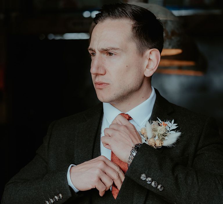 The groom in a black suit with an orange tie wearing a dried flower and bunny grass buttonhole for the rustic glam wedding 