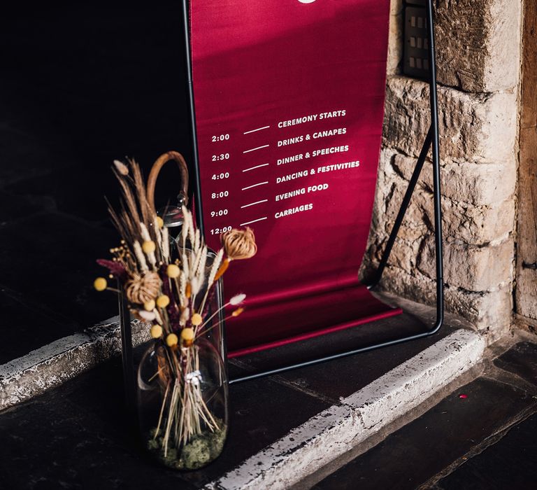 Red fabric order of the day wedding sign on black metal frame for autumnal boho wedding with dried grass in vase 