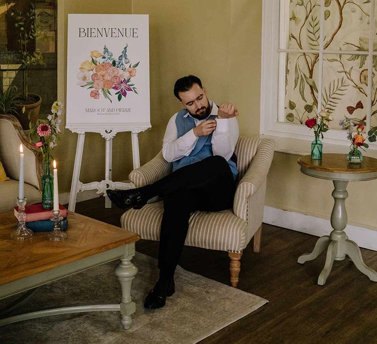 Groom wearing cornflower blue waistcoat and black suit trousers fixing his shirt in the reception room of Braxted Park wedding venue 