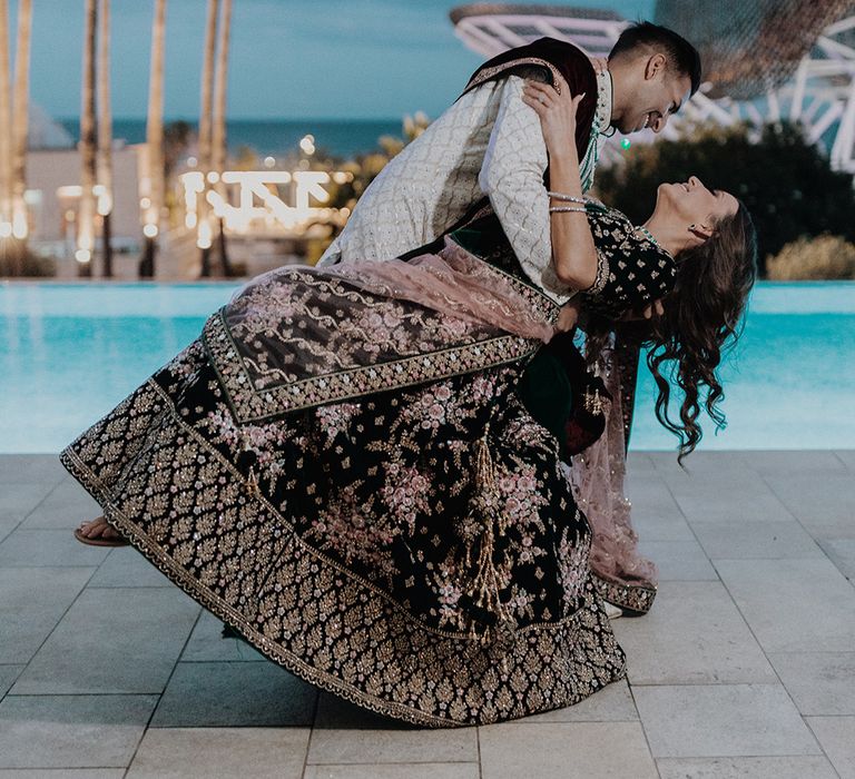 Groom in white and gold patterned sherwani by the pool at castle wedding venue in Barcelona dipping bride in two piece black and gold patterned saree dancing