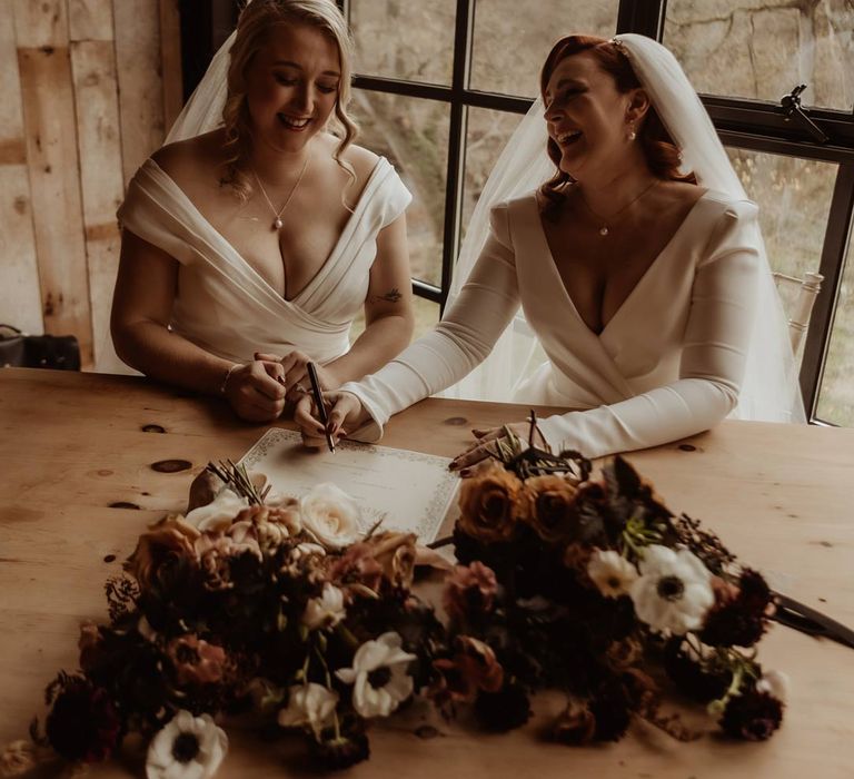 Brides in complimenting deep v wedding dress signing the register at Hidden River Cabins after LGBTQIA wedding ceremony with white, dusky pink and burnt red dried flower, rose and peony bridal bouquets