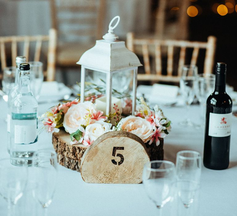 A table centrepiece with a white lantern and ring of florals with a table number sign on a tree trunk 