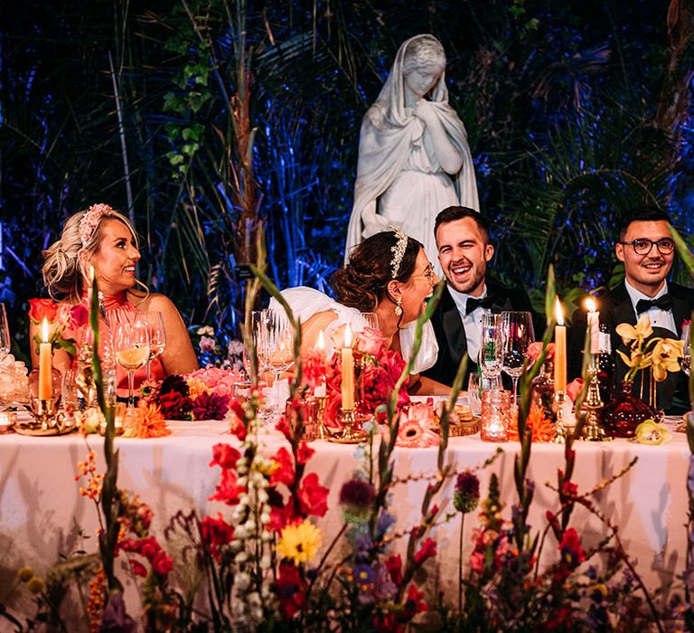 The bride and groom laugh together sitting at their colourful top table decorated with bright flowers 