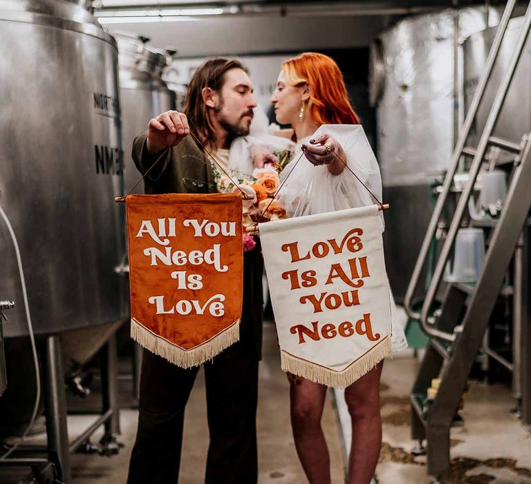 Bride and groom holding up velvet fabric wedding banners with retro lettering in burnt orange and white standing by beer fermenters at Northern Monk Refectory Leeds