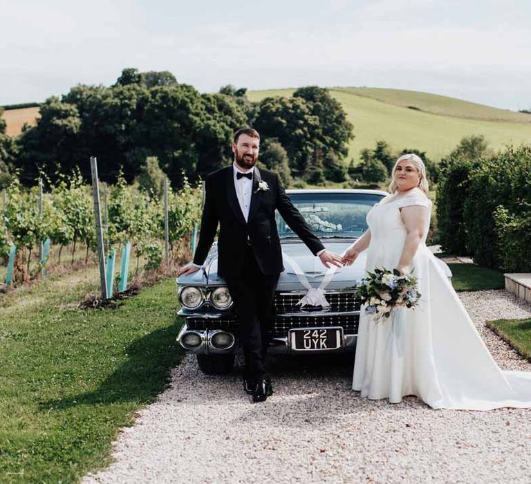 Groom in classic black grooms three piece tuxedo with white rose boutonniere and black bowtie holding hands with bride in satin off the shoulder wedding dress holding white and blue eucalyptus, foliage, garden rose, gardenia, light blue delphinium and dried flower bridal bouquet tied with dusky blue ribbon standing in front of baby blue classic wedding car 