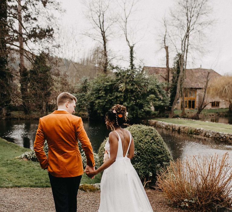 Bride in low back sleeveless wedding dress with 3D floral applique train walking with groom in crushed burnt orange velvet grooms blazer