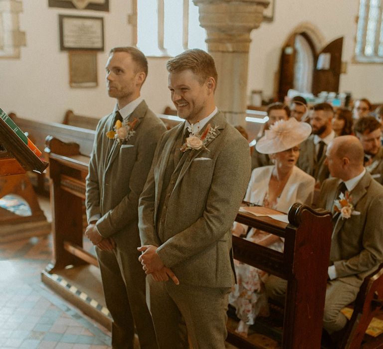 The groom and best man stand waiting at the aisle for the bride at their church wedding 