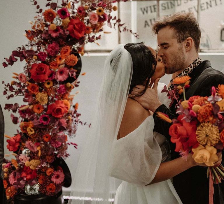 Bride in off shoulder tulle short wedding dress with church length veil kissing groom wearing wet look black grooms suit with leopard print shirt and pink and orange boutonniere under vinyl wedding arch