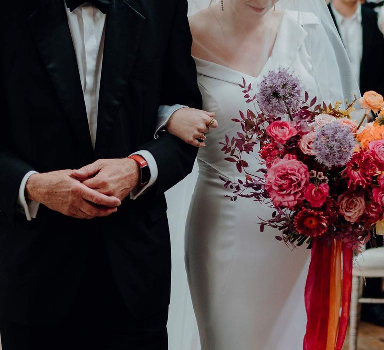 Father of the bride walking down the aisle with bride in asymmetrical off shoulder long sleeve satin wedding dress with cathedral length personalised veil holding colourful bridal bouquet with pink garden roses, purple giant alliums, dark red poppies and foliage 