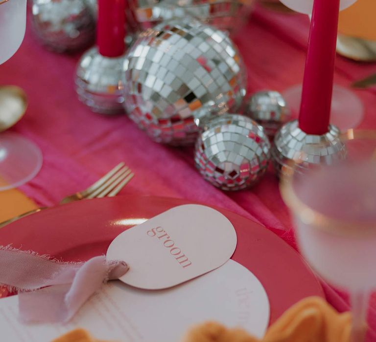 Circular pink wedding place name on pink and orange wedding tablescape with bright pink wedding tablecloth, orange wedding napkins, pink tapered candles, pink and orange poppies, garden roses, carnations, dried flowers and foliage wedding table centrepieces and disco ball wedding decor 