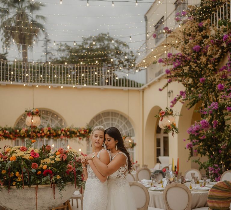 two bride in lace wedding dresses embracing at their outdoor wedding reception at a Croatia wedding venue with string lights 