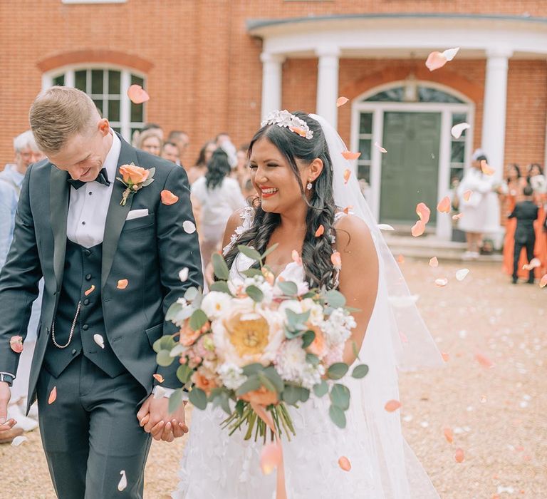 Bride in floral lace princess wedding dress with groom in black tuxedo walking out of their bespoke ceremony to confetti 