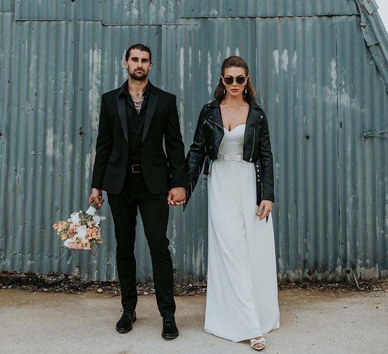 Groom in full black wedding suit standing with bride in strapless bow wedding dress with leather jacket and black sunglasses 