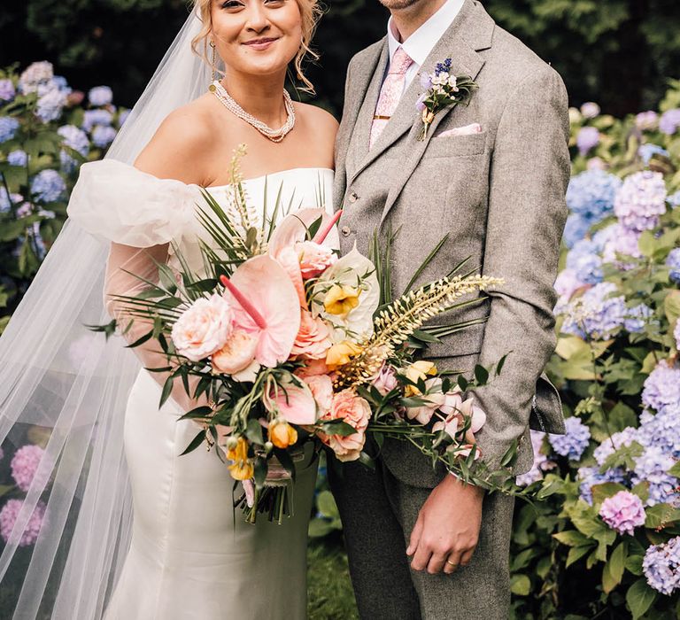 Bride wears an off the shoulder puff sleeve wedding gown holding a tropical wedding bouquet with the groom in tweed suit 