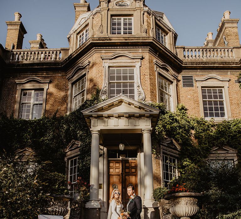 The bride wears a long sleeve wedding dress with retro sunglasses next to the groom on the steps on Hampton Court House wedding venue 