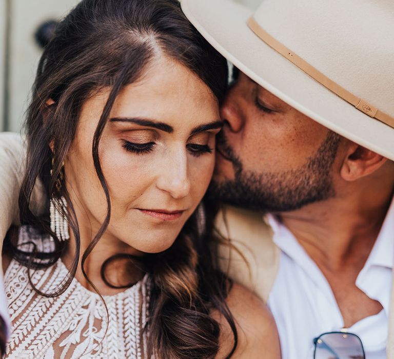 Bride with ponytail hairstyle being kissed on the cheek by the groom in a hat 