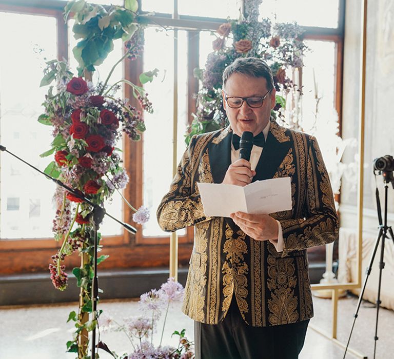 Groom in black and gold patterned Dolce & Gabbana suit jacket reading vows at wedding ceremony 