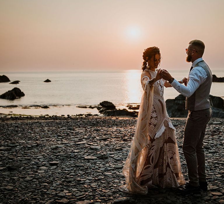 Sunset golden hour wedding photo with the bride in her lace gown at the beach with the groom in a grey suit 