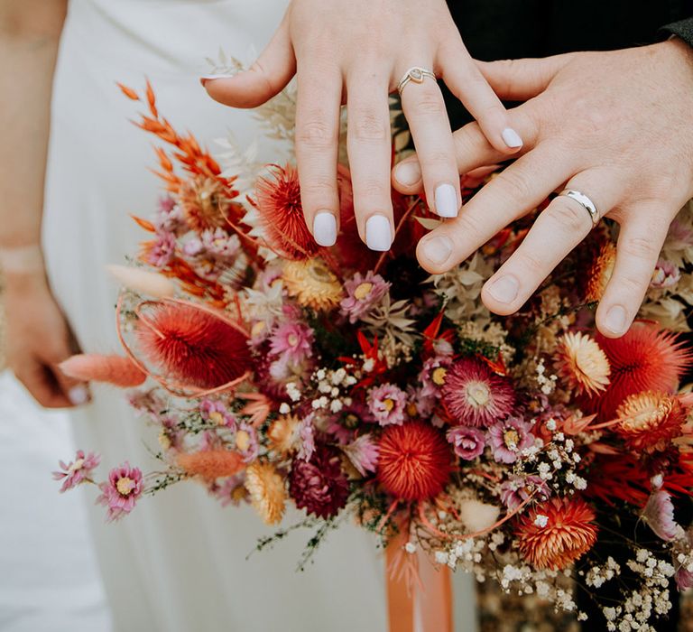 Bride carrying a pink, red and white wedding bouquet for her summer wedding showing off the new wedding rings with the groom 