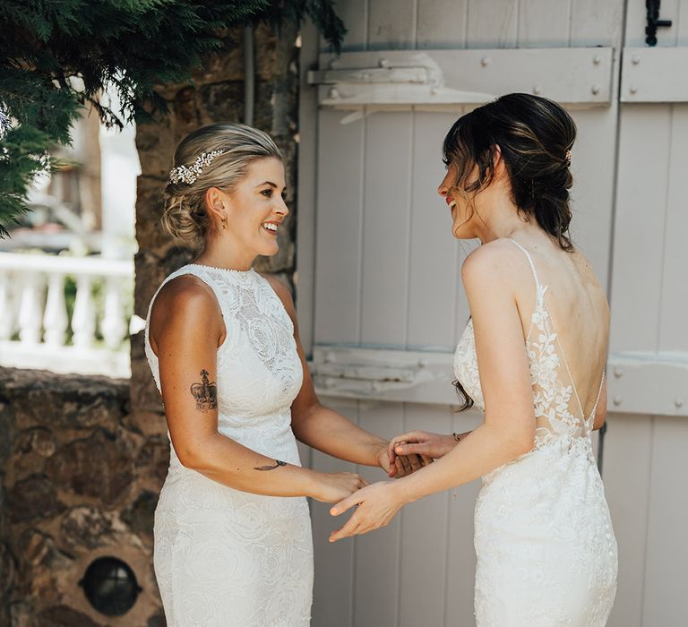 Two brides in lace wedding dresses holding hands as they have a first look moment between each other 