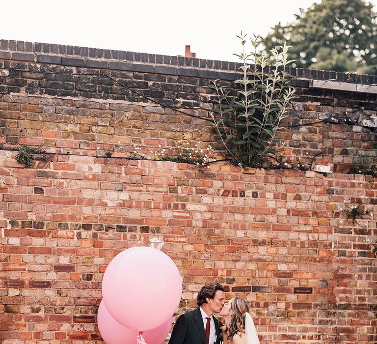 Bride in strapless fitted wedding dress posing with groom in dark green wedding suit with large pink wedding balloons