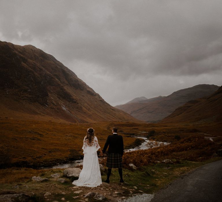 Bride and groom stand hand in hand looking at the highlands in Glencoe