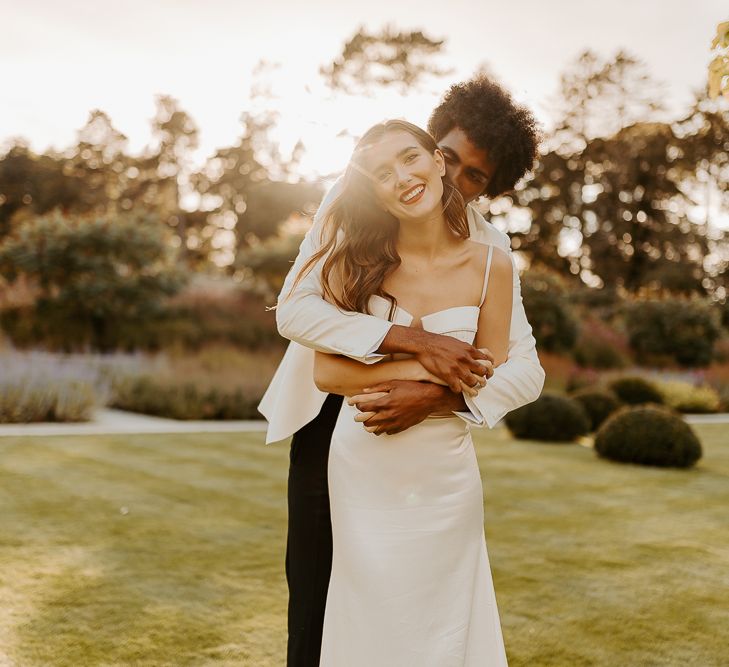 Golden hour portrait with the Black groom with afro hair and white tuxedo jacket embracing his bride with long brown hair and red lipstick